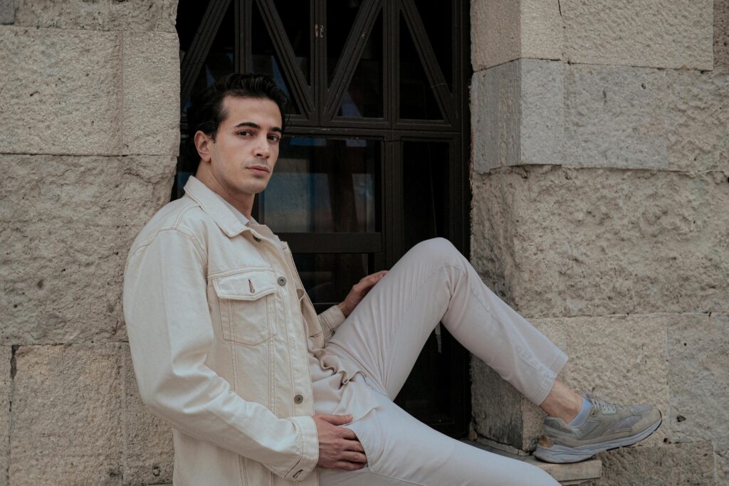 Young man in cream jacket and white pants posing against stone wall, urban fashion.