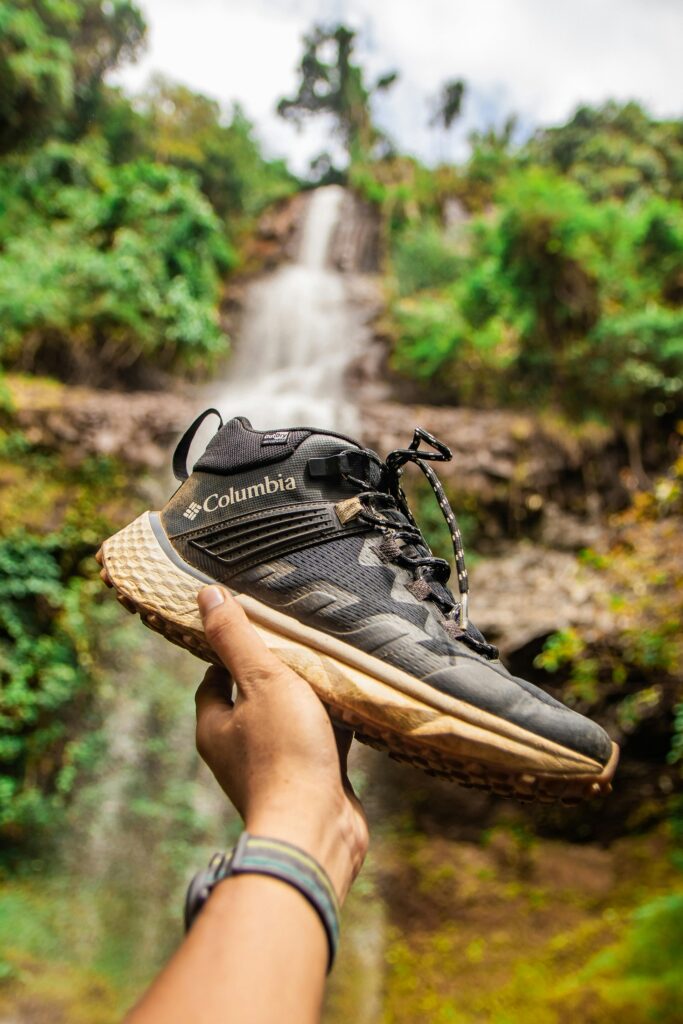 A Columbia shoe held in front of a majestic waterfall in Đắk Nông, Việt Nam, showcasing adventure.